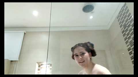 Media: Video of a fair-skinned woman with dark hair styled in twin buns, smiling in a modern, beige-tiled bathroom with mosaic accents and a glass shower door.