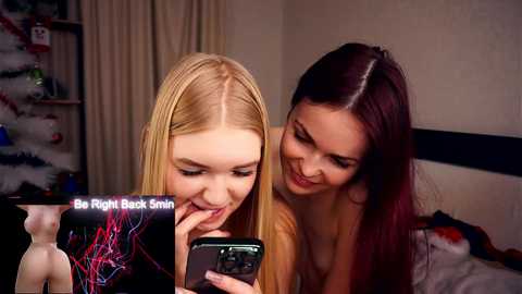 Video of two young women, one blonde, one brunette, playing a video game on a smartphone. They are in a cozy bedroom with a bed and shelves in the background.