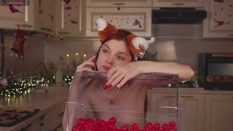 Media: Video of a pale-skinned, red-haired woman in a kitchen with a festive, holiday-themed background. She leans on a countertop, wearing a sheer, see-through top and a pair of fox ears headband.