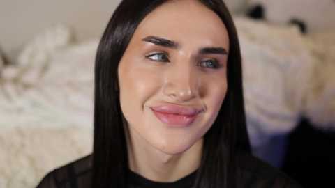 Media: Video of a smiling, fair-skinned woman with straight, long black hair, wearing black clothing, against a blurred background featuring white bedding.