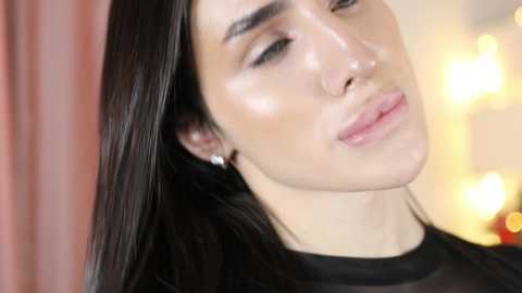 Media: Video of a fair-skinned transgender woman with long, straight black hair, wearing a black shirt, and a subtle makeup look with soft pink lips. Background shows blurred, warm-toned lights.