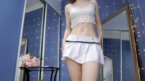 Media: Video of a slender, light-skinned woman with long brown hair, wearing a white lace bralette and mini skirt, standing in a blue bedroom with floral arrangements and mirrors.