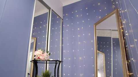 Media: Video of a room with lavender walls adorned with white twinkling fairy lights and a large mirror with a gold frame. A black stand with pink flowers sits in the foreground.