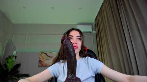 Media: Video of a young woman with long brown hair, wearing a light blue t-shirt, holding a large, dark, veiny dildo, in a modern, beige-toned room with curtains and a painting on the wall.