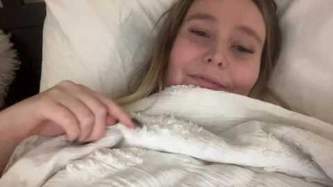 Media: A video of a young Caucasian woman with blonde hair, lying in bed, partially covered by a white blanket. She has a content expression, and her hand is touching the blanket. The background is blurry, indicating a cozy, indoor setting.