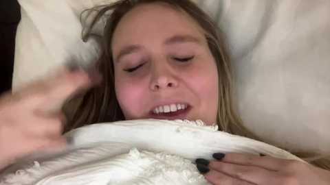 Media: Video of a young woman with fair skin, light brown hair, and closed eyes, lying on white pillows. She has black nail polish and a slight smile.