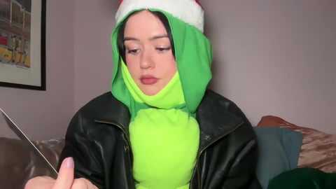 Media: Video of a young woman with fair skin, wearing a neon green hooded jacket over a black leather jacket, and a Santa hat. She is seated indoors, holding a cigarette, with a framed picture on the wall in the background.