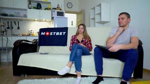 Media: Video of a modern kitchen with a woman in a red plaid shirt and jeans, and a man in a grey t-shirt and blue jeans, both sitting on a cream couch.