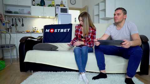 Media: Video of a man and woman on a beige couch in a modern kitchen. Man wears a gray t-shirt, woman a red plaid shirt.