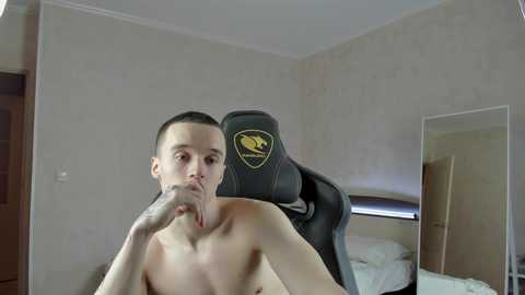 Media: A video of a shirtless, light-skinned man with short dark hair, wearing a black gaming chair with a yellow logo, sitting in a minimalist bedroom with beige walls, wooden door, and a mirror.