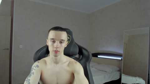 Media: Video of a young, fair-skinned, shirtless man with short hair, tattoos on his right arm, sitting on a black gaming chair in a minimalist bedroom with beige walls and a bed in the background.