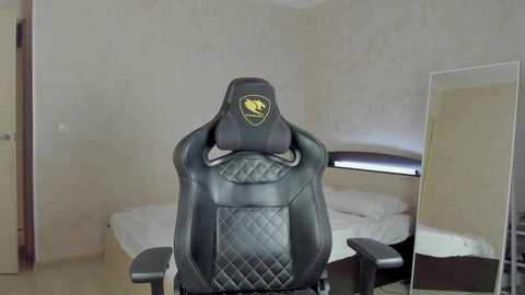 Media: Video of a black, quilted, ergonomic gaming chair with a built-in headrest featuring a yellow and black logo in a modern, minimalist bedroom with a bed, mirror, and light-colored walls.