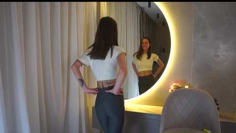 Media: A video of a woman with long dark hair, wearing a white crop top and high-waisted jeans, standing before a round mirror with a yellow light.