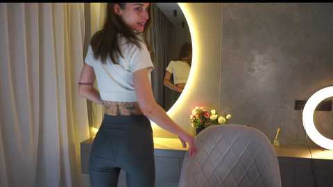 Media: Video of a young woman with medium-length brown hair, wearing a white crop top and high-waisted gray pants, adjusting a cushion in a modern, minimalist bedroom with a circular mirror and a glowing lamp.