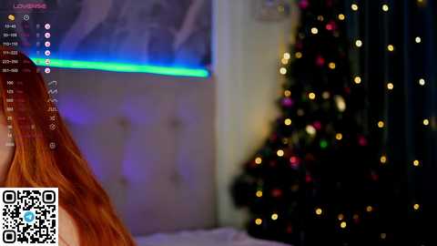 Media: Video of a woman with long red hair, seated at a desk, in a dimly lit room with a decorated Christmas tree and a glowing LED strip in the background.