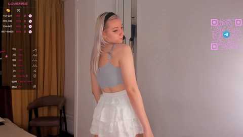 Media: Video of a blonde woman with a slender figure, wearing a light blue crop top and white ruffled skirt, standing in a modern, dimly lit room with a TV displaying a game interface.