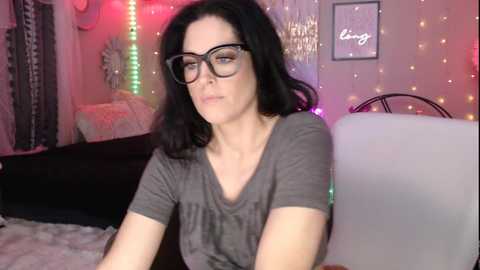 Media: Video of a middle-aged woman with black hair and glasses, wearing a gray t-shirt, seated on a bed with pink fairy lights, a TV, and a white pillow.