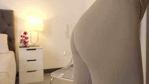 Media: Video of a person's buttocks in tight beige leggings, standing beside a white nightstand with a lamp and red flowers.