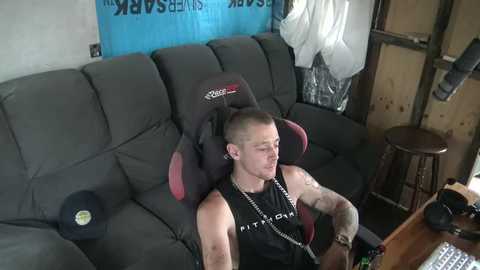 Media: A video of a muscular man with tattoos, wearing a black sleeveless shirt, seated in a dark grey sectional sofa, gaming with a headset, in a cluttered, dimly lit room.