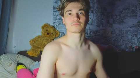 Media: Video of a young, shirtless, slim, white male with short, light brown hair, sitting in a dimly lit room with a blue and white wallpaper. A yellow teddy bear is visible on a bed behind him.