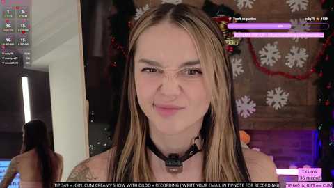 Media: Video of a young woman with light skin, long blonde hair, and a black choker, smiling in a festive room with holiday decorations, including snowflakes. Text overlays indicate streaming activity.