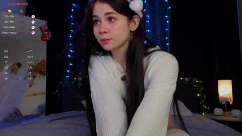 Video of a young woman with long dark hair, wearing a cream knit sweater, in a cozy, dimly lit room with twinkling blue fairy lights, a lit candle, and a bed in the background.