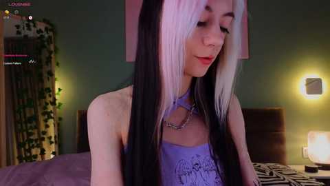 Media: Video of a slender, light-skinned transgender woman with long, straight black and white hair. She wears a purple halter top, sitting on a bed with green walls and a bedside lamp.