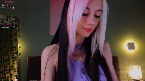 Video of a young woman with long black and white hair, wearing a light purple lace top, smiling, in a dimly lit bedroom with green walls and a wall-mounted lamp.