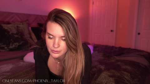 Media: A video of a young woman with long, straight, blonde hair, wearing a black top, leaning on a bed with a dark, patterned comforter in a dimly lit room with pinkish-orange lighting.