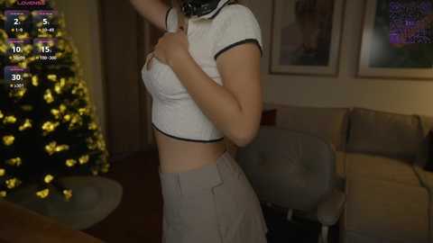 Media: Video of a young woman with light skin, wearing a white crop top and grey skirt, standing in a dimly lit room with a decorated Christmas tree and a plush couch.