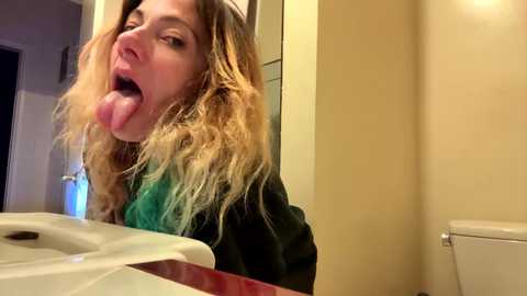 Media: A candid video of a young woman with long, wavy blonde hair sticking out of a white toilet seat, tongue hanging out, wearing a green shirt and black jacket.