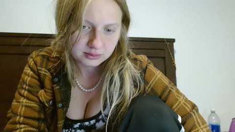 Media: A video of a young Caucasian woman with long, wavy blonde hair, wearing a brown plaid shirt, black top, and pearl necklace, sitting on a bed, with a dark-haired man's head resting on her shoulder.