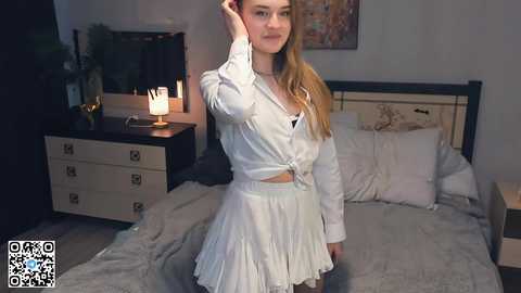 Media: A video of a young Caucasian woman with long blonde hair, wearing a white, ruffled crop top and skirt, standing in a dimly-lit bedroom with a bed, dresser, and potted plant.