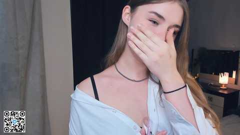 Media: Video of a young Caucasian woman with long, light brown hair, wearing a white shirt, black bra, and a choker, covering her mouth with her hand. Bedroom background with candles, bed, and dark curtains.