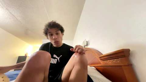 Media: A video of a young man with curly hair, wearing a black Puma shirt, sitting on a wooden bed in a dimly lit room, reaching for a foot.