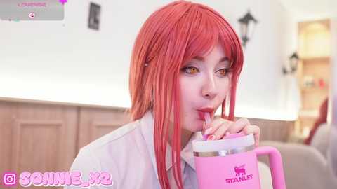 Media: Video of a young woman with straight, long, vibrant red hair and fair skin, wearing a white shirt, drinking from a pink Stanley's cup, in a modern kitchen with light wooden cabinets.