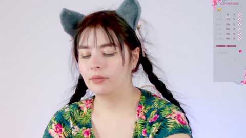 Media: A video of a young woman with long black hair, wearing cat ears and a floral shirt, eyes closed, standing against a white wall.