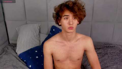 Media: Video of a young, shirtless, slim, light-skinned man with curly brown hair, seated on a bed with a grey blanket, white and striped pillows, and a blue pillow with white stars.