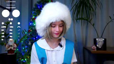 Video of a young woman with short blonde hair wearing a blue and white dress, a fluffy white hat, and a microphone. She stands next to a decorated Christmas tree with blue lights, a potted plant, and a wooden table in the background.