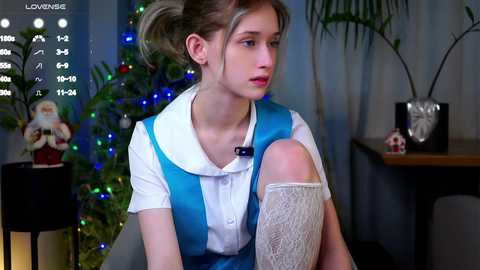 Media: A video of a young woman with light skin, brown hair tied back, wearing a blue and white dress, sitting with a bandaged leg, in a Christmas-decorated room with a potted plant and a Santa figure.