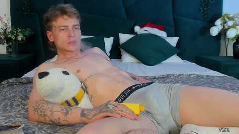 Media: Video of a fit, tattooed young man in gray Calvin Klein underwear, lying on a bed with a panda plush, green headboard, white pillows, and Christmas decorations.