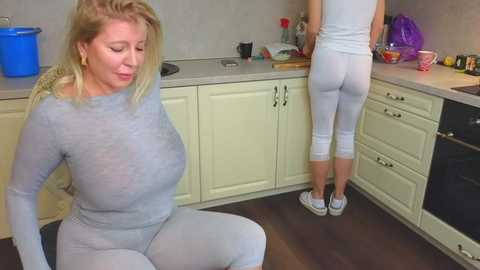 Media: Video of a blonde woman in a tight, gray workout outfit, sitting on a kitchen floor. Behind her, a woman in white leggings and a tank top stands near a counter.