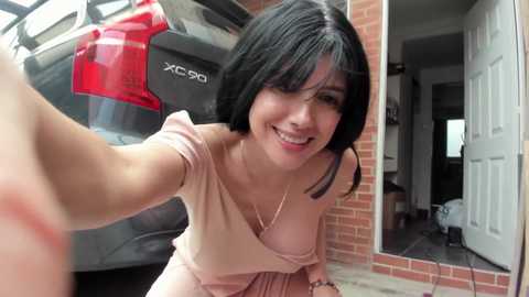 A video of a smiling, fair-skinned woman with short black hair in a pink dress, kneeling beside a parked SUV, with a brick wall and doorway visible in the background.