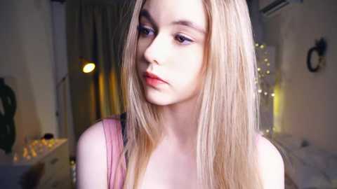 Media: Video of a young Caucasian woman with long, straight blonde hair, fair skin, and red lipstick, wearing a pink tank top, standing in a dimly lit bedroom with a green lamp and fairy lights in the background.