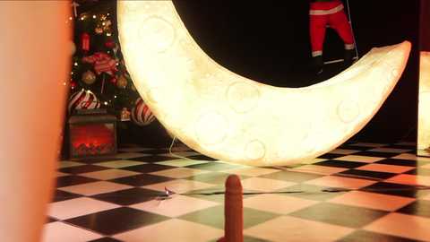 Media: Video of a festive room with a glowing, moon-shaped light on a black-and-white checkered floor. A Christmas tree with ornaments is partially visible in the background. A person in red pants and a black top stands in the shadows.