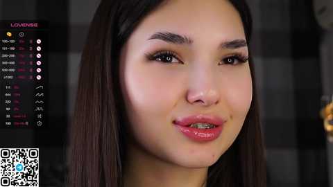 Video of a young Asian woman with long, straight black hair and fair skin. She has sharp eyebrows, small round nose, and full lips. The background is blurred with a checkered pattern.