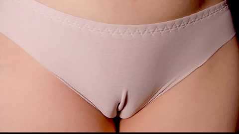 Media: Close-up video of a person's lower abdomen and upper thighs, showing beige, smooth, elasticized panties with a scalloped waistband. The skin is fair and smooth, and the lighting is soft.