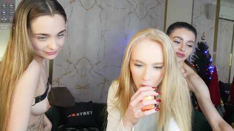 Media: Video of three young women: one blonde with a white top, another with long brown hair in a black bra, and a third with dark hair, all indoors, drinking.