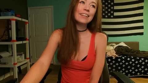 Media: Video of a smiling young woman with long brown hair, wearing a red tank top and black choker, sitting in a cluttered bedroom with a star-patterned wall and bookshelf.