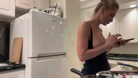 Media: Video of a blonde woman in a dark tank top, checking her phone, standing in a modern kitchen with white cabinets and a black countertop.